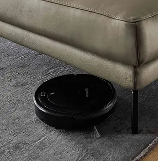 Saugroboter reinigt unter einem modernen olivgrünen Ledersofa mit schwarzen Metallfüßen. Detailansicht auf Teppichboden, zeigt hohe Bodenfreiheit und praktische Alltagstauglichkeit des Sofas.